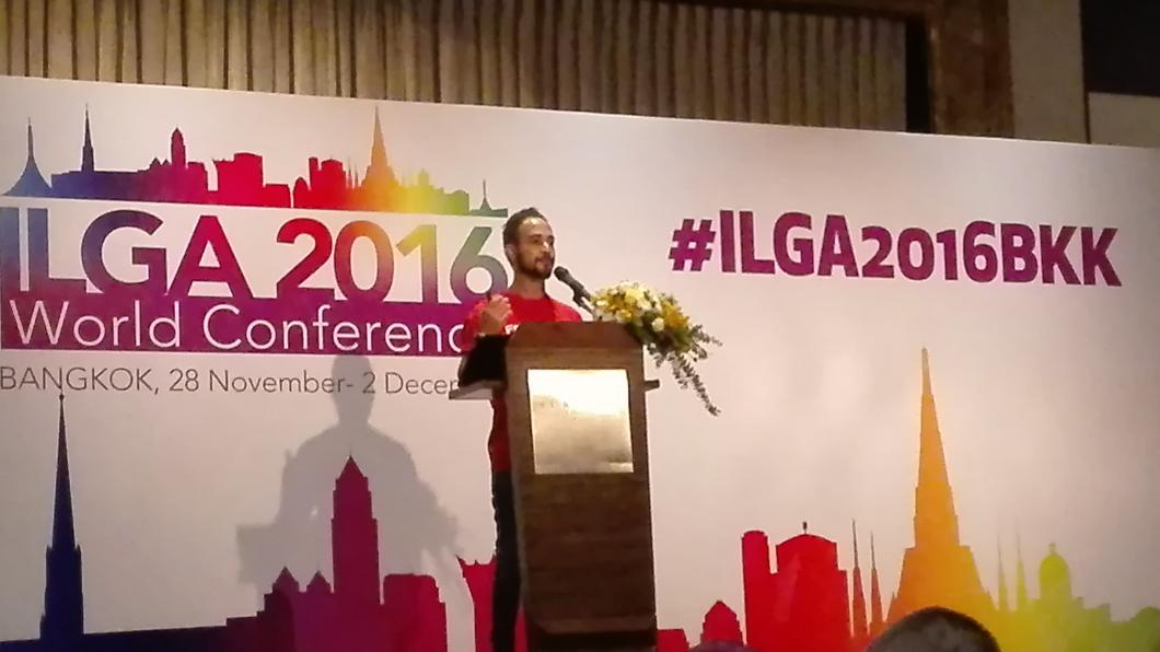 Spreker op spreekgestoelte op podium; daarachter spandoeken met ILGA2016BKK (de afkorting voor Bangkok)